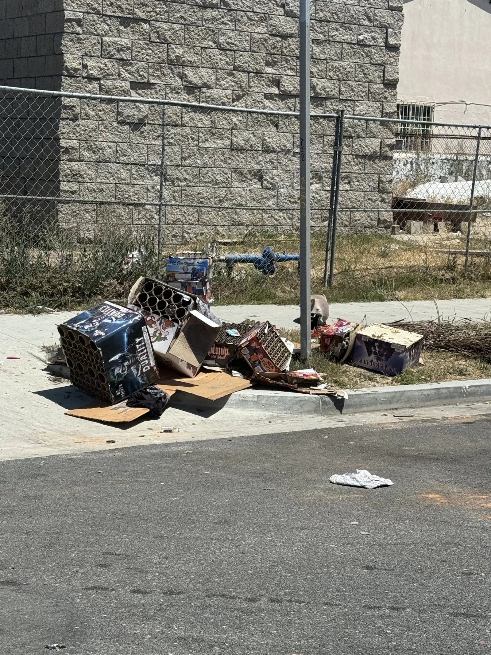 A large pile of flattened boxes, cardboard packaging, tubes, and other debris is visible along the sidewalk and curb beside a chain-link fence.